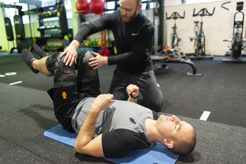 A man performs a core stability exercise on a gym floor with the help of a sports therapist